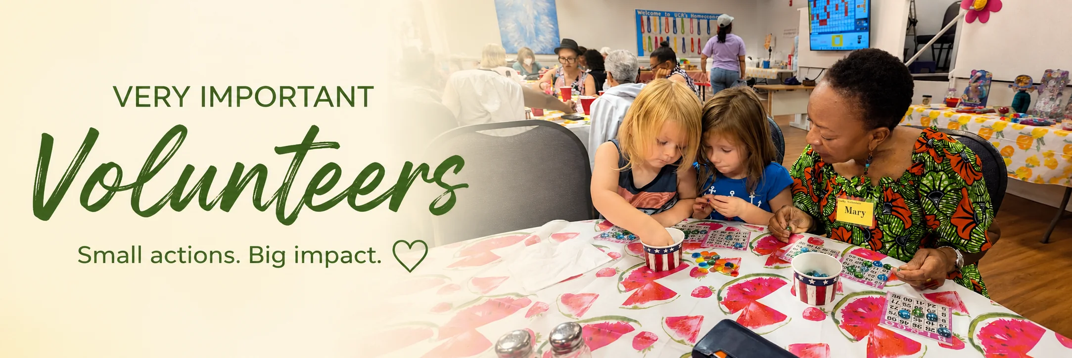 A Unity of Raleigh volunteer wearing a name badge sitting with two young children at a bingo table, helping them place their markers, with a homecoming gathering going on behind them. Very Important Volunteers — small actions, big impact.